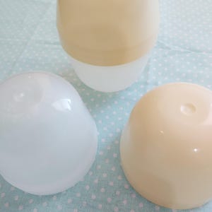 Może przedstawiać: Trzy szklane przedmioty w odcieniach bieli i beżu ułożone są na jasnoniebieskiej powierzchni w kropki. Największy obiekt ma dwukolorowy kształt jaja. Widoczne są również dwa mniejsze, zaokrąglone obiekty.