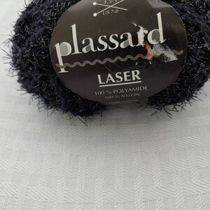 Könnte beinhalten: Ein schwarzer Wollknäuel mit einem weißen Etikett, auf dem "plassard LASER 100% POLYAMIDE 100% NYLON" steht. Das Etikett enthält auch das Garngewicht, die Nadelstärke und die Lauflänge.