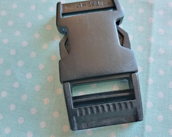 May include: A grey plastic buckle, marked with "A1-29B", sits on a light blue fabric dotted with white polka dots. The buckle is rectangular, featuring slots and a locking mechanism, commonly used in various applications.