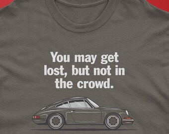 May include: A charcoal gray crew neck t-shirt featuring a white graphic of a vintage sports car in profile. Above the car, the text reads, "You may get lost, but not in the crowd." The design is centered on the chest of the shirt.