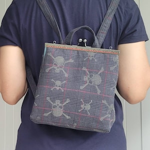 Könnte beinhalten: Ein grauer und schwarzer karierter Rucksack mit einem silbernen Verschluss und einem Totenkopfdesign. Der Rucksack hat einen blauen und roten Besatz.