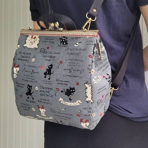 May include: A gray canvas backpack with black cat illustrations and white text. The backpack has a flap closure with a gold metal clasp. The backpack is being worn by a person.