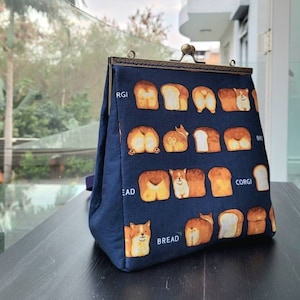Puede incluir: Un bolso de mano de tela azul marino con un cierre de latón. El bolso tiene un patrón repetido de traseros de perro corgi y rebanadas de pan. El texto "Bread", "Corgi" y "RGI" es visible en el bolso.