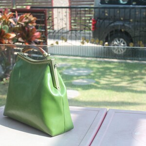 May include: A green leather handbag with a rectangular shape and a clasp closure. The bag has a wooden handle and gold-colored metal accents. The bag is sitting on a pink surface.