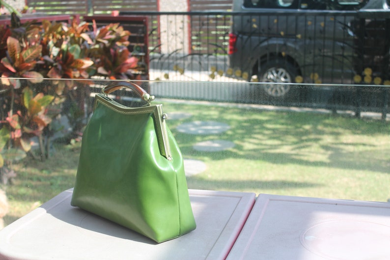 May include: A green leather handbag with a rectangular shape and a clasp closure. The bag has a wooden handle and gold-colored metal accents. The bag is sitting on a pink surface.