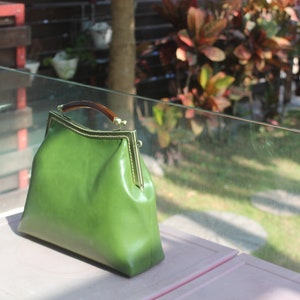 May include: A green leather handbag with a gold-colored frame and a tortoiseshell handle. The bag is sitting on a pink surface, with a blurred outdoor background.