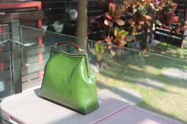 May include: A green leather handbag with a gold-colored frame and a tortoiseshell handle. The bag is sitting on a pink surface, with a blurred outdoor background.
