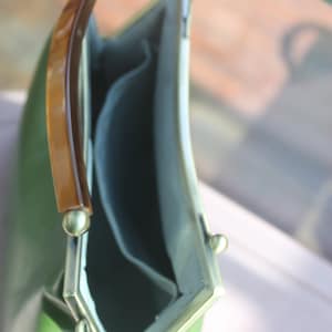 May include: A vintage-style green handbag with a curved shape and a tortoiseshell handle. The bag has a light green exterior, a light blue interior, and gold-colored metal accents. The clasp is open, revealing the interior.