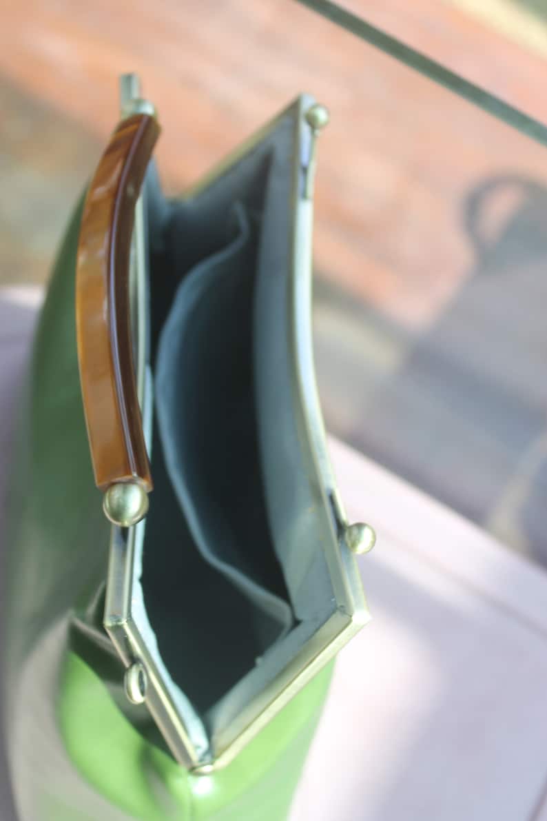 May include: A vintage-style green handbag with a curved shape and a tortoiseshell handle. The bag has a light green exterior, a light blue interior, and gold-colored metal accents. The clasp is open, revealing the interior.