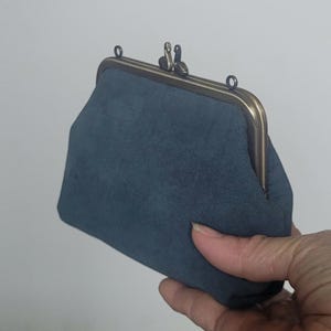 May include: A slate blue pouch with a soft, matte texture and a bronze-toned metal frame. The closure features two small, sculpted bird-shaped knobs at the top, and the frame includes two small circular loops for attaching a strap or chain.