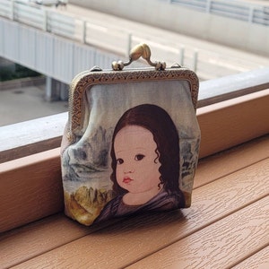 Puede incluir: Un pequeño bolso de estilo vintage con un cierre de latón y un diseño de tela que presenta un retrato de una niña con el pelo largo y oscuro. El bolso está hecho de una tela suave y texturizada y tiene un fondo azul claro.