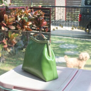 May include: A green leather handbag with a gold-colored clasp and a brown handle. The bag is sitting on a pink surface, with a blurred background of a yard and a small dog.