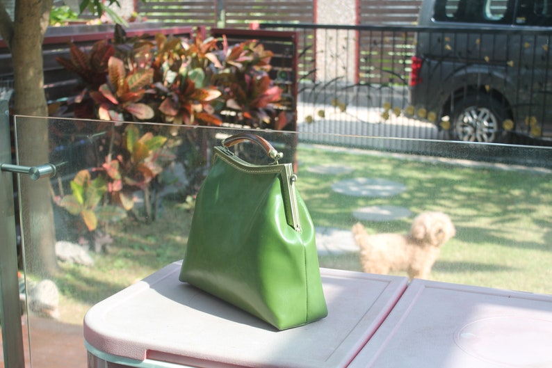 May include: A green leather handbag with a gold-colored clasp and a brown handle. The bag is sitting on a pink surface, with a blurred background of a yard and a small dog.