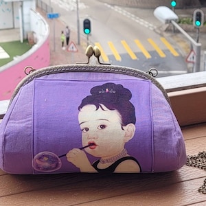 Puede incluir: Un bolso de mano de tela morado con cierre de metal y correa de cadena. El bolso presenta una imagen impresa de una niña pequeña soplando una burbuja con una pajita.