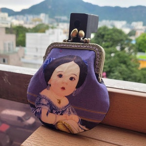 Puede incluir: Una cartera de monedas de tela morada con un cierre de estilo vintage y una imagen impresa de una niña con ojos azules y un vestido morado.
