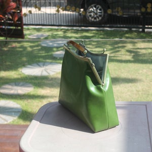 May include: A green handbag with a gold-colored frame and a wooden handle. The bag is open, revealing a gray interior. The bag is sitting on a light-colored surface, with a green lawn in the background.