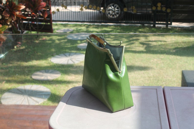May include: A green handbag with a gold-colored frame and a wooden handle. The bag is open, revealing a gray interior. The bag is sitting on a light-colored surface, with a green lawn in the background.