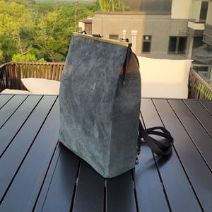 以下が含まれることがあります： 金属製のフレームと黒いストラップが付いた青と茶色のスエードのバックパック。バックパックは木製のテーブルの上に置かれています。