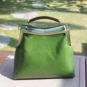 May include: A green leather handbag with a metal frame and a brown handle. The bag has a rectangular shape with a rounded top and a clasp closure. The bag is sitting on a white surface.