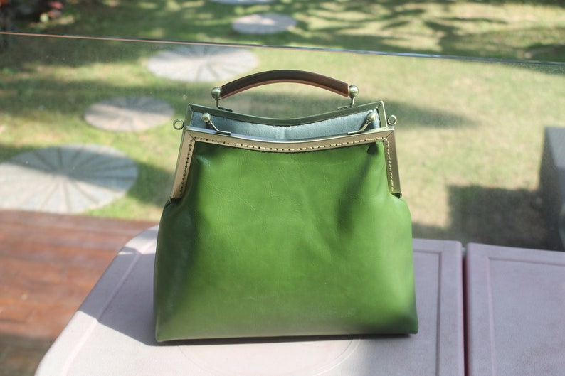May include: A green leather handbag with a metal frame and a brown handle. The bag has a rectangular shape with a rounded top and a clasp closure. The bag is sitting on a white surface.