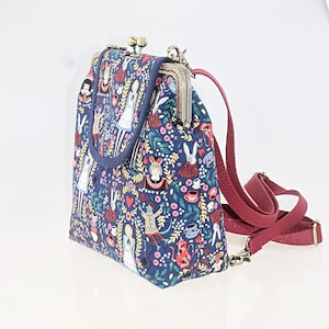 May include: A blue canvas backpack with a floral print featuring Alice in Wonderland characters. The bag has a metal clasp closure and an adjustable burgundy strap.