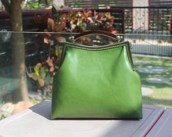 Puede incluir: Un bolso de mano de cuero verde con un asa marrón. El bolso tiene una forma estructurada con un cierre metálico y un ribete dorado. El bolso está sobre una superficie blanca.