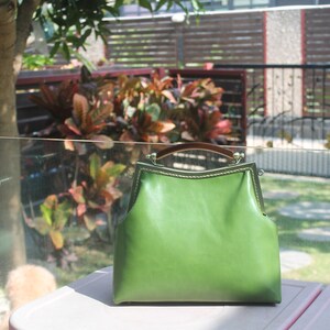 May include: A green leather handbag with a structured frame and a brown handle. The bag has a clasp closure and gold-colored metal accents. The bag is sitting on a pink surface.