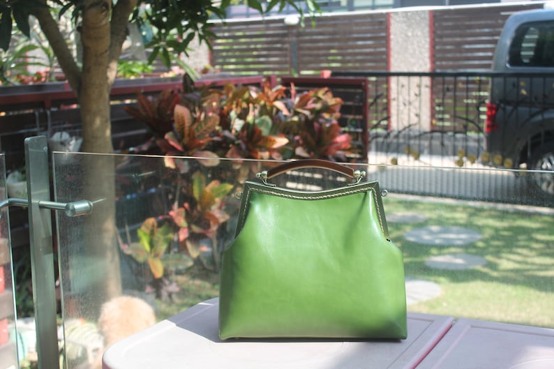 May include: A green leather handbag with a structured frame and a brown handle. The bag has a clasp closure and gold-colored metal accents. The bag is sitting on a pink surface.