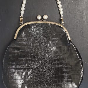 Könnte beinhalten: Schwarze, ovale Handtasche mit einer Textur aus Kunstkrokodilleder. Die Tasche hat einen perlenbesetzten Griff und einen goldfarbenen Verschluss. Zwei perlenartige Perlen schmücken die Oberseite des Verschlusses. Der Hintergrund ist dunkel.