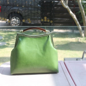 May include: A green leather handbag with a brown handle and a metal clasp. The bag has a structured shape and a vintage aesthetic. The bag is sitting on a white surface.