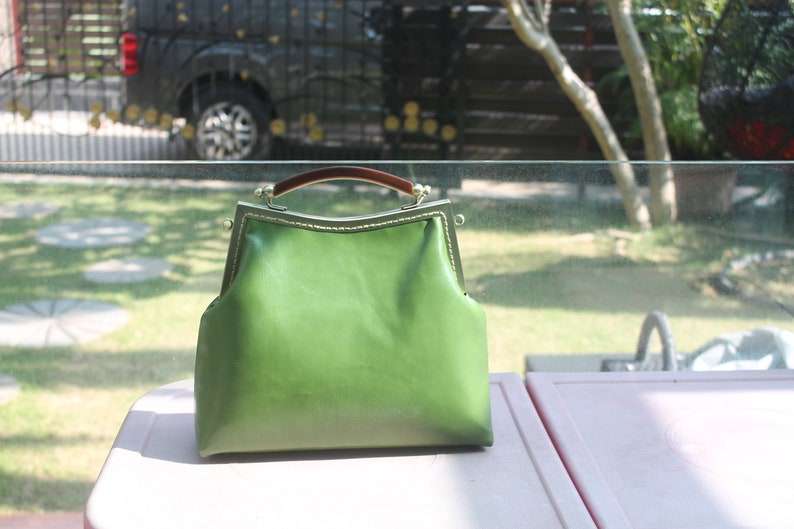 May include: A green leather handbag with a brown handle and a metal clasp. The bag has a structured shape and a vintage aesthetic. The bag is sitting on a white surface.