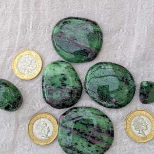 May include: Five polished, smooth, oval-shaped green and dark red stones are arranged alongside three gold-coloured coins. The stones feature mottled patterns of deep forest green with patches of dark magenta or burgundy throughout the surfaces.