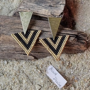 Könnte beinhalten: Ein Paar goldene und schwarze Dreiecksohrringe mit geometrischem Muster. Die Ohrringe sind aus Perlen gefertigt und haben ein modernes Design.