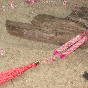 May include: A pink and white beaded necklace with a red tassel. The necklace is made with small beads and has a silver chain. The tassel is attached to the end of the necklace.