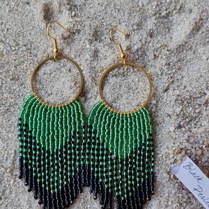 May include: A pair of green and black beaded hoop earrings with gold hoops and fringe. The earrings are on a sandy surface with a small white tag that says "Black Pearls".