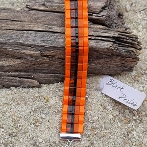 Peut inclure: Bracelet en perles orange et noires avec un fermoir argenté. Le bracelet est composé de petites perles carrées et porte une étiquette indiquant "Black Perles".