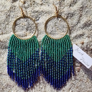Peut inclure: Une paire de boucles d'oreilles à franges perlées avec des cerceaux dorés. Les boucles d'oreilles présentent un dégradé de perles vertes et bleues, avec une étiquette indiquant "Black Perles". Les boucles d'oreilles sont présentées sur une surface sablonneuse.