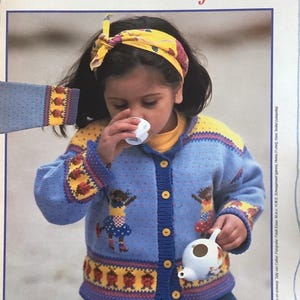 May include: A child wearing a blue cardigan sweater with a mouse design, yellow accents, and a matching headband. The sweater has a button-up front and is paired with a small white teapot. The text "het Vera de Muis-vestje" is at the top.