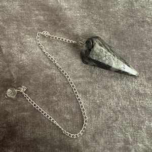 May include: A pointed, faceted pendulum made of a black and white marbled stone, attached to a silver-toned metal chain. The chain features a small, clear, spherical bead at the end, providing a balanced weight for the pendulum's movement.