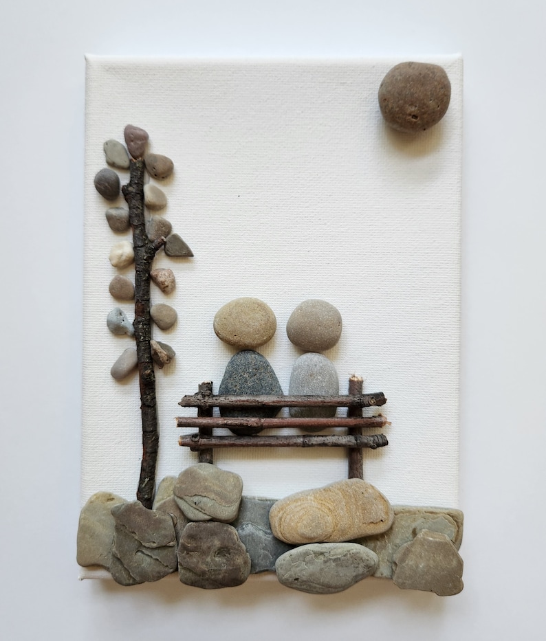 May include: A stone art piece depicting two figures sitting on a bench made of twigs. The figures are made of gray and brown stones. A small tree made of twigs and stones is on the left side of the image. The background is a white canvas.