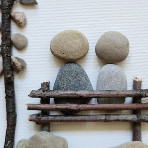 May include: Two gray and brown stones are arranged to look like people sitting on a bench made of small branches. The bench is made of four branches, and the stones are sitting on the top two branches.