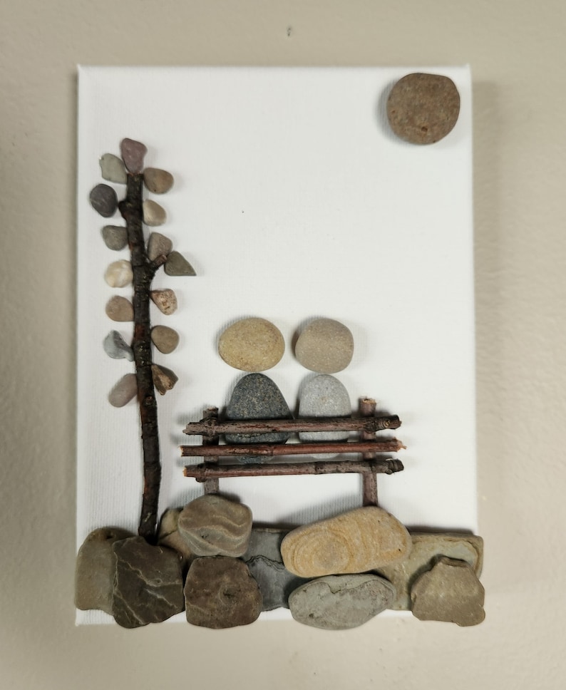 May include: A white canvas with a stone art piece depicting two figures sitting on a bench made of twigs. The figures are made of stones and the background includes a stone tree and a stone sun.