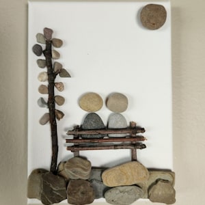 May include: A white canvas with a stone art piece depicting two figures sitting on a bench made of twigs. The figures are made of stones and the background includes a stone tree and a stone sun.
