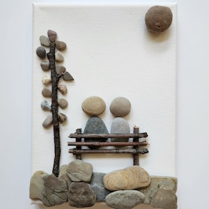 May include: A stone art piece depicting two figures sitting on a bench made of twigs. The figures are made of gray and brown stones. A small tree made of twigs and stones is on the left side of the image. The background is a white canvas.