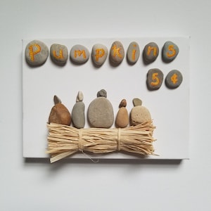 May include: A white canvas with the word "Pumpkins" spelled out in orange painted stones.  Five smaller stones are arranged in a row below the word, with the number "5" and a dollar sign painted on them.  A row of gray and brown stones are arranged in a row below the dollar sign, with a bundle of straw in front of them.