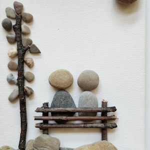 May include: A stone sculpture of two figures sitting on a bench made of twigs. The sculpture is on a white background with a small tree made of stones and twigs to the left.