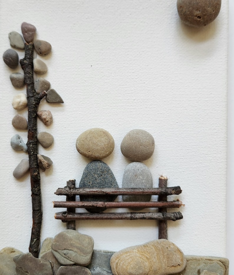 May include: A stone sculpture of two figures sitting on a bench made of twigs. The sculpture is on a white background with a small tree made of stones and twigs to the left.