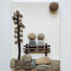 May include: A stone art piece depicting two figures sitting on a bench made of twigs. The figures are made of gray and brown stones. A small tree made of twigs and stones is on the left side of the image. The background is a white canvas.