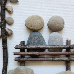 May include: Two gray and brown stones are arranged to look like people sitting on a bench made of small branches. The bench is made of four branches, and the stones are sitting on the top two branches.