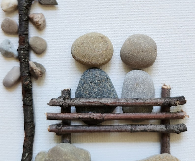 May include: Two gray and brown stones are arranged to look like people sitting on a bench made of small branches. The bench is made of four branches, and the stones are sitting on the top two branches.